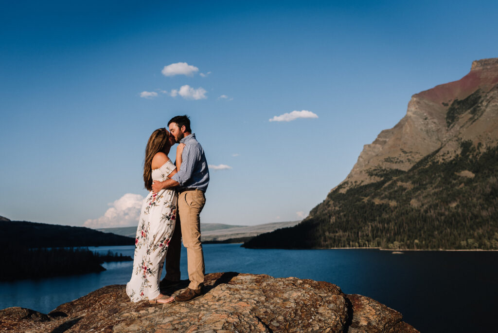 man and woman kissing on top of rock overlooking a lake with mountains in the background at Glacier National Park, Montana