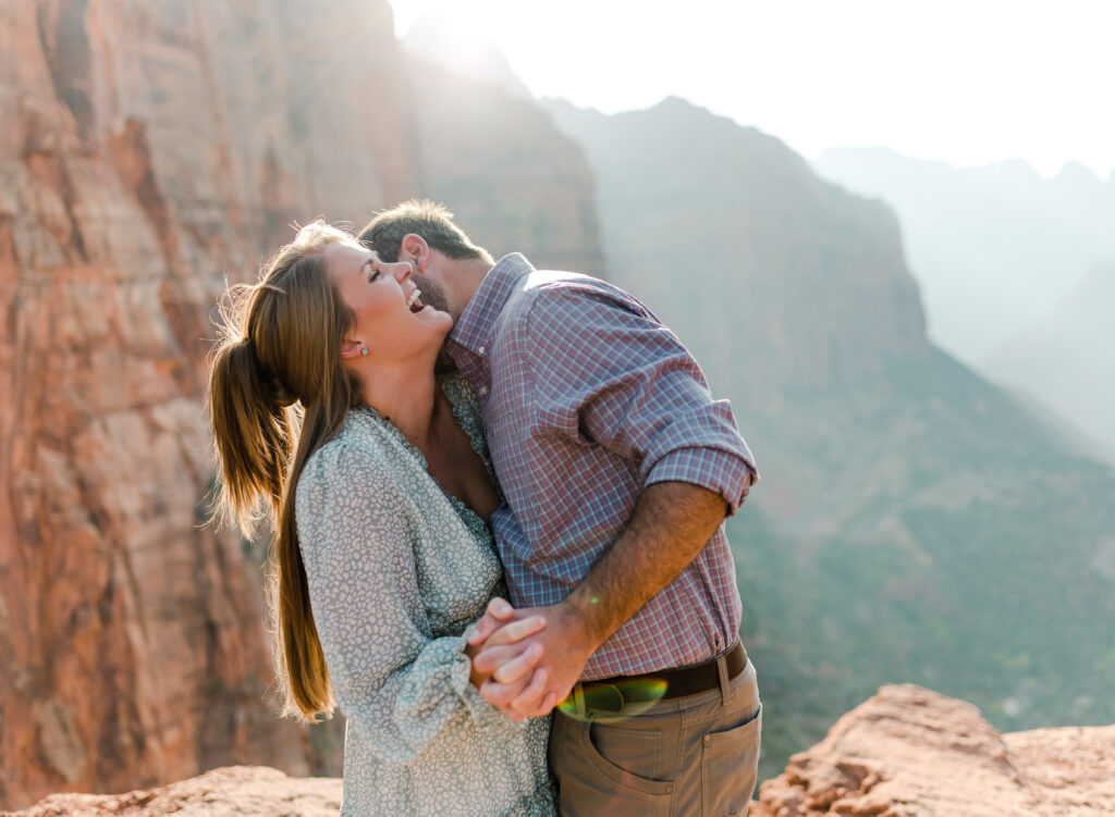 A man kisses wife while she laughs while on a lookout in Zion National Park at their adventure elopement.