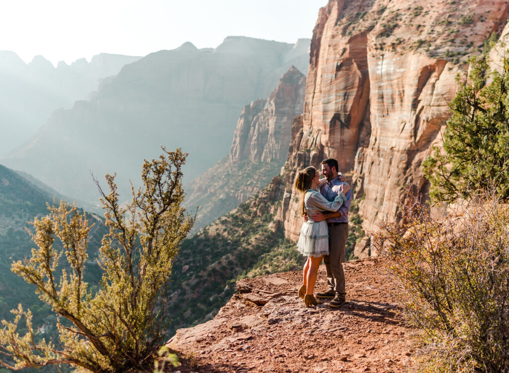 A man and woman on adventure elopement embrace cliffside at Zion National Park with stunning mountain landscape backdrop