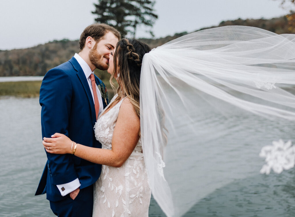 A bride and groom kiss on their elopement day as her veil flows behind her in the air. Background of a lake.