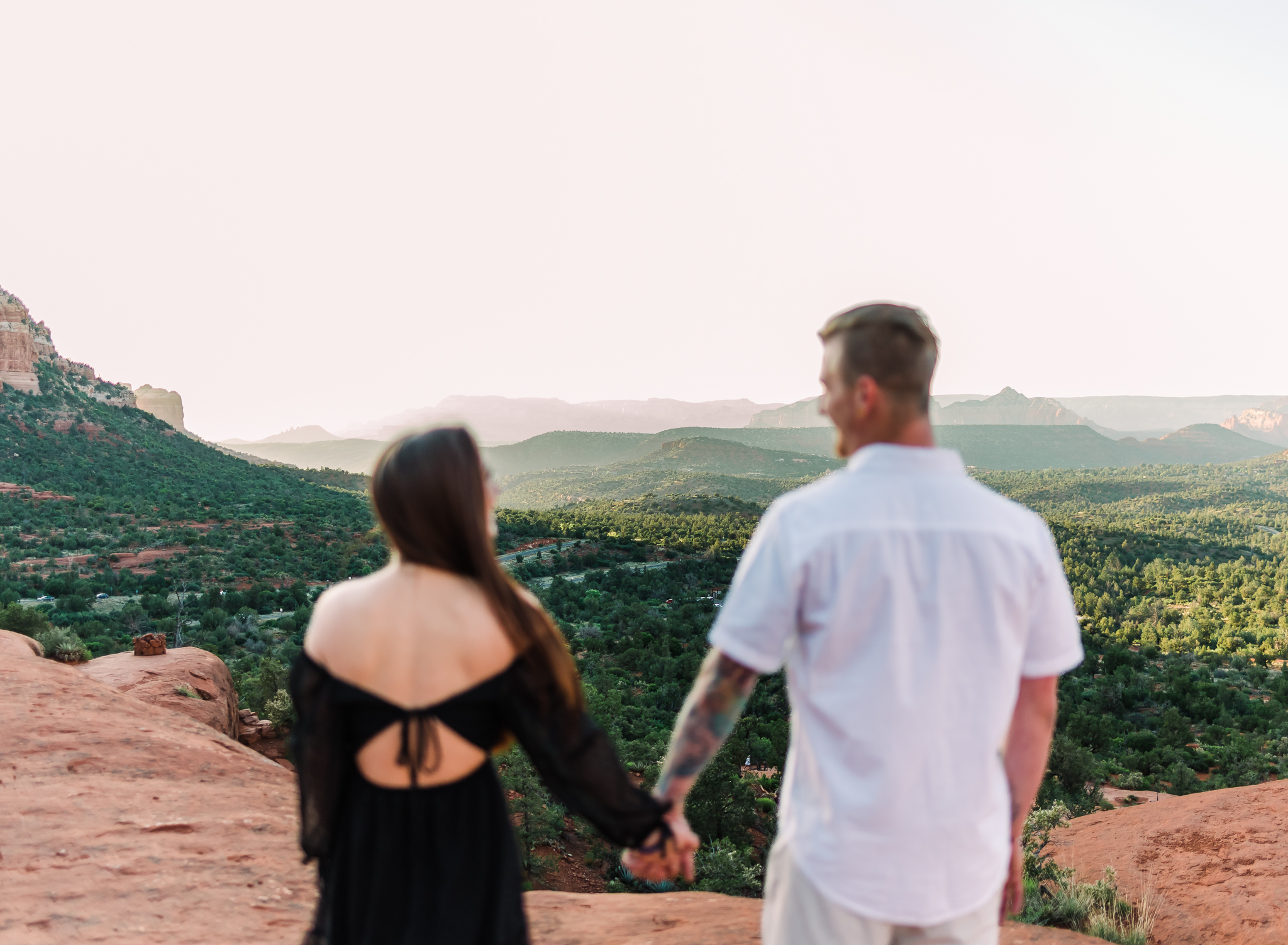 A man and a woman hold hands and look at each other. THey are out of focus while the landscape in front of them is in focus. The landscape is the red rocks of Sedona Arizona