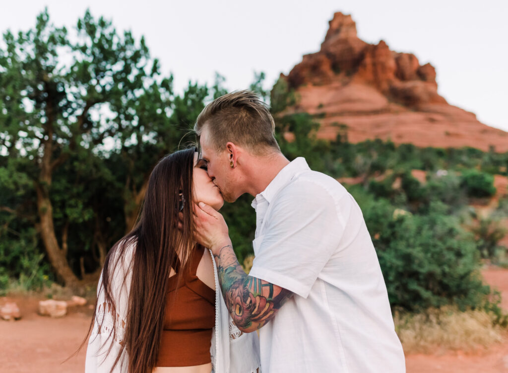 A man and woman kiss with Bell Rock visible behind them in Sedona Arizona
