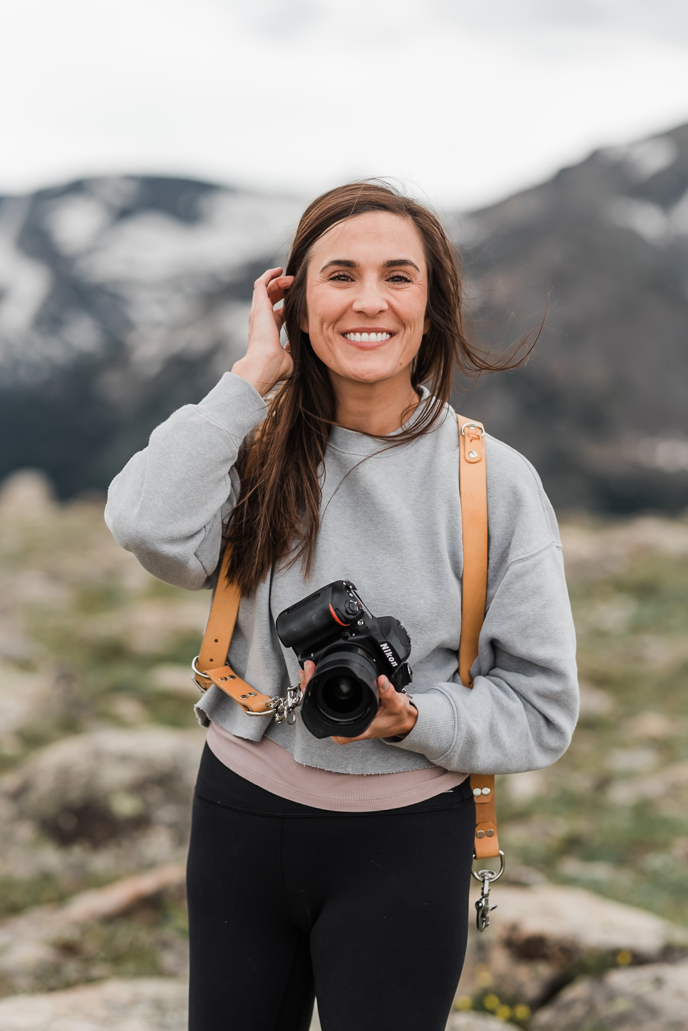 A woman holding her camera in one hand and putting her hair behind her ear with the other hand. She is smiling at the camera and this is a headshot for her photography business