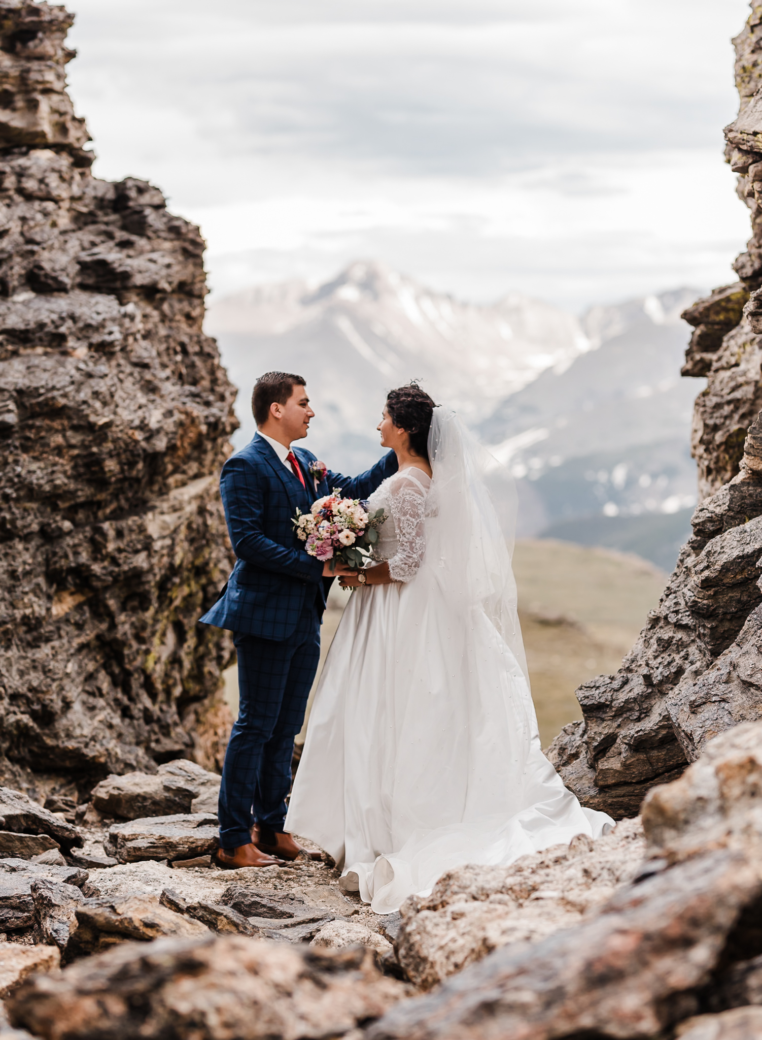 A bride and groom are standing on top of rocks with large mountains behind them while they exchange vows