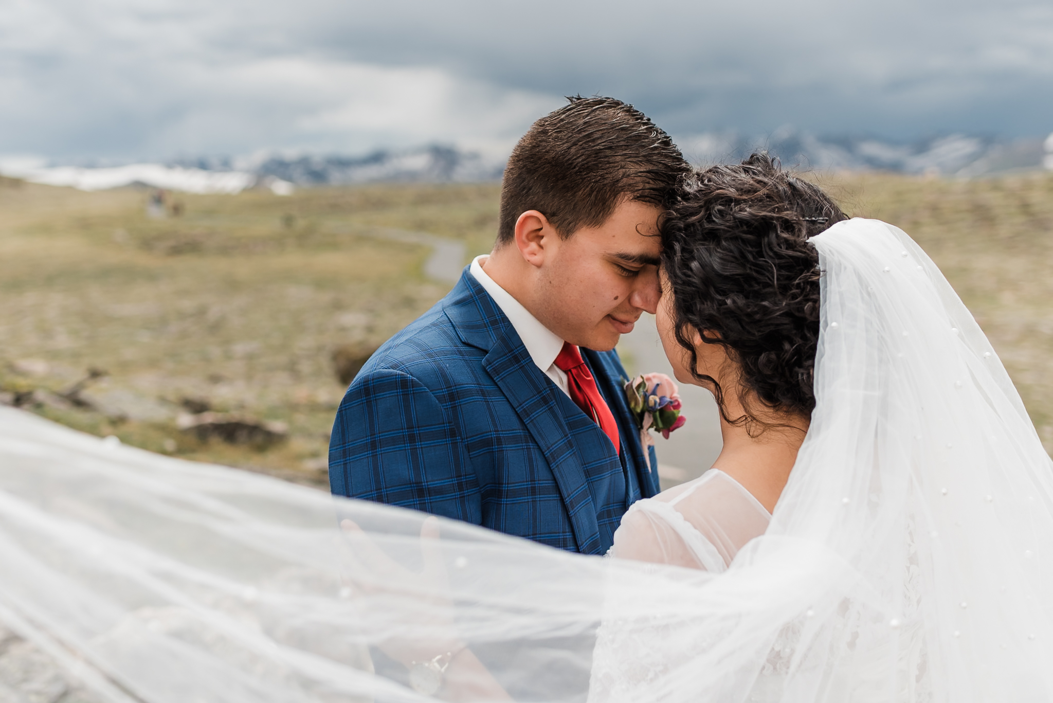 A bride and groom are touching foreheads and looking into each others eyes, while the brides veil blows in the wind. There are mountains behind them