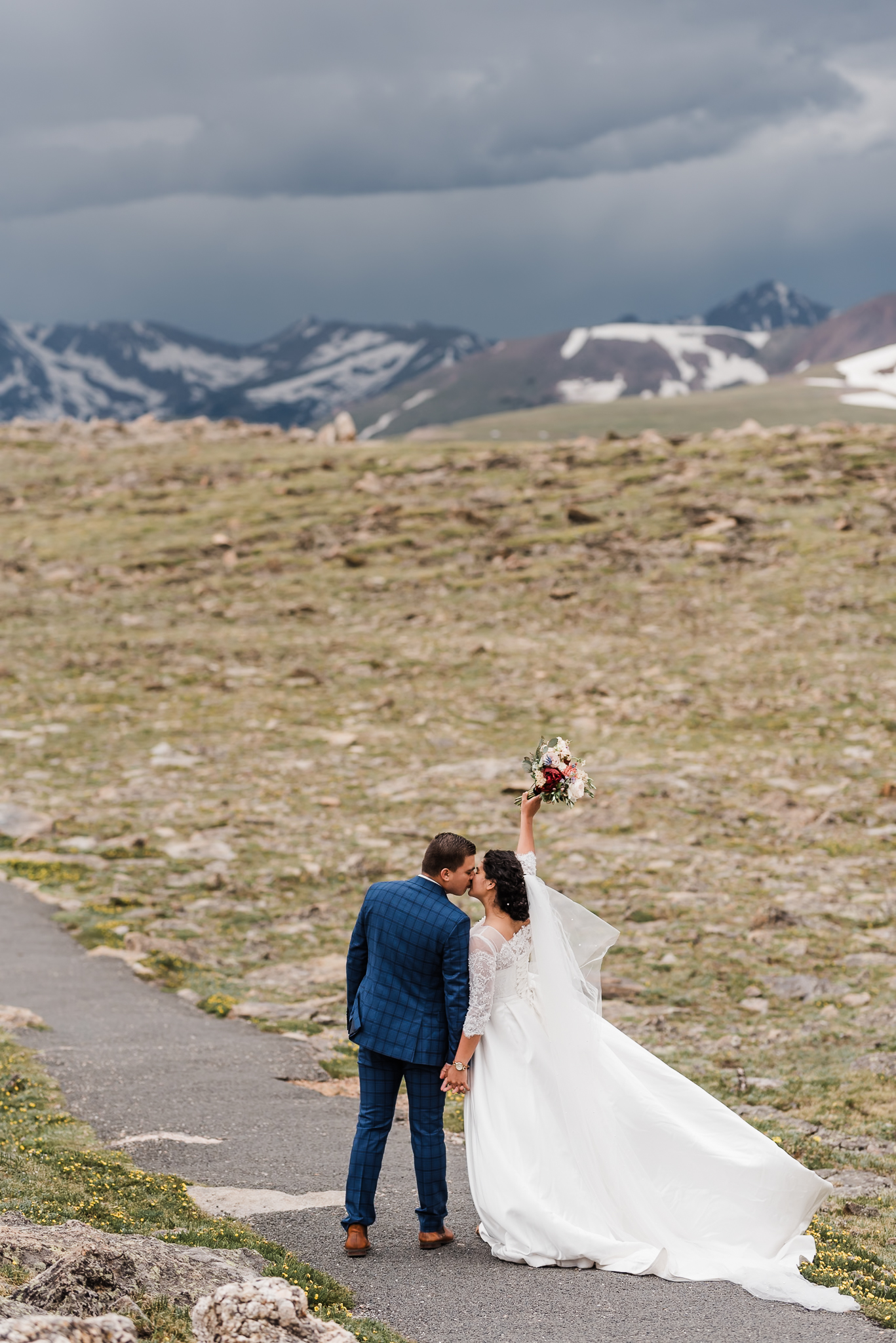 A bride and groom kiss and the bride holds her bouquet up in celebration of their marriage. There is an open field and large mountains behind them at their colorado elopement