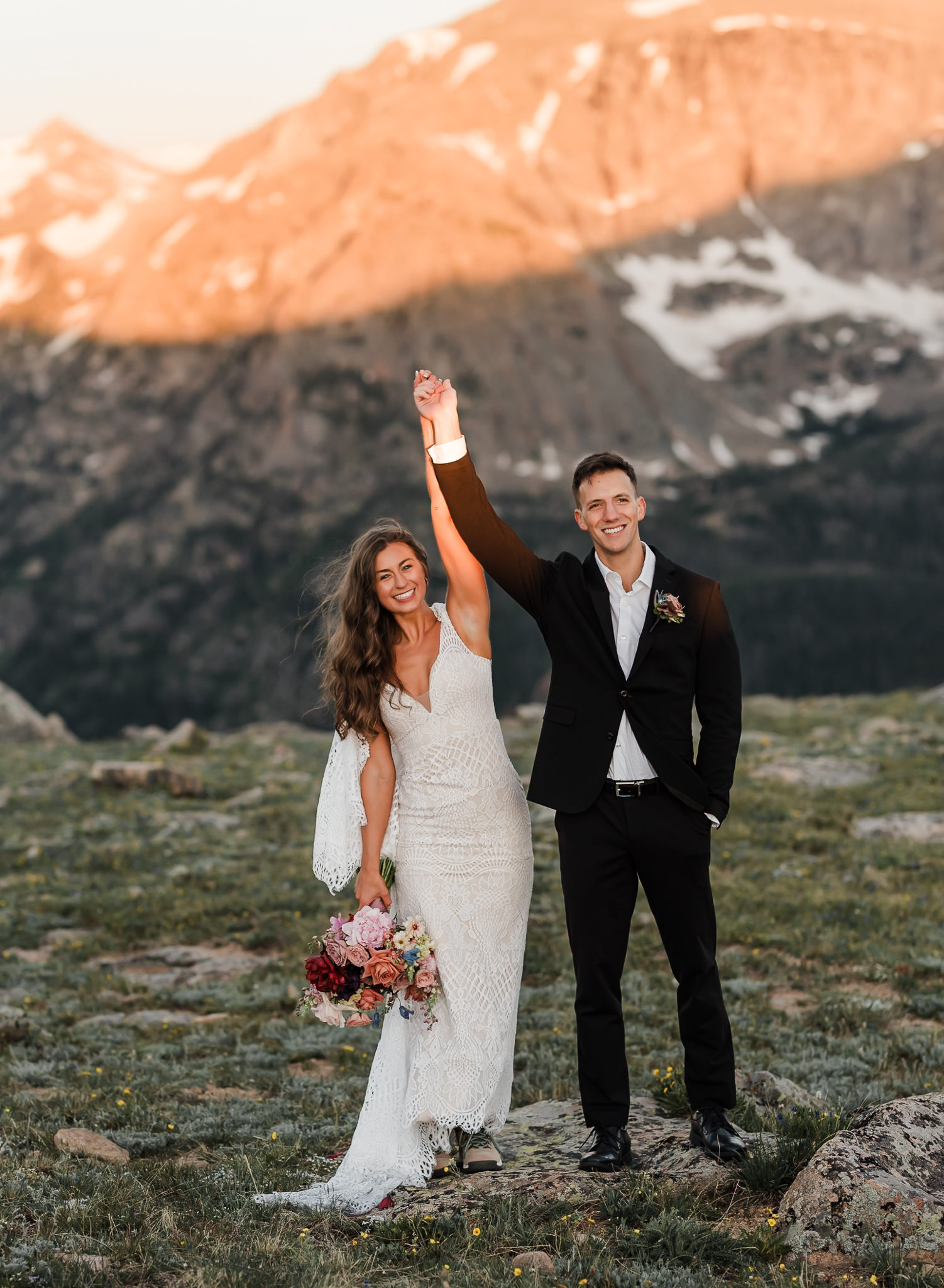 A bride and groom are holding hands and raising them in celebration after exchanging wedding vows in front of a large mountain at Rocky Mountain National Park