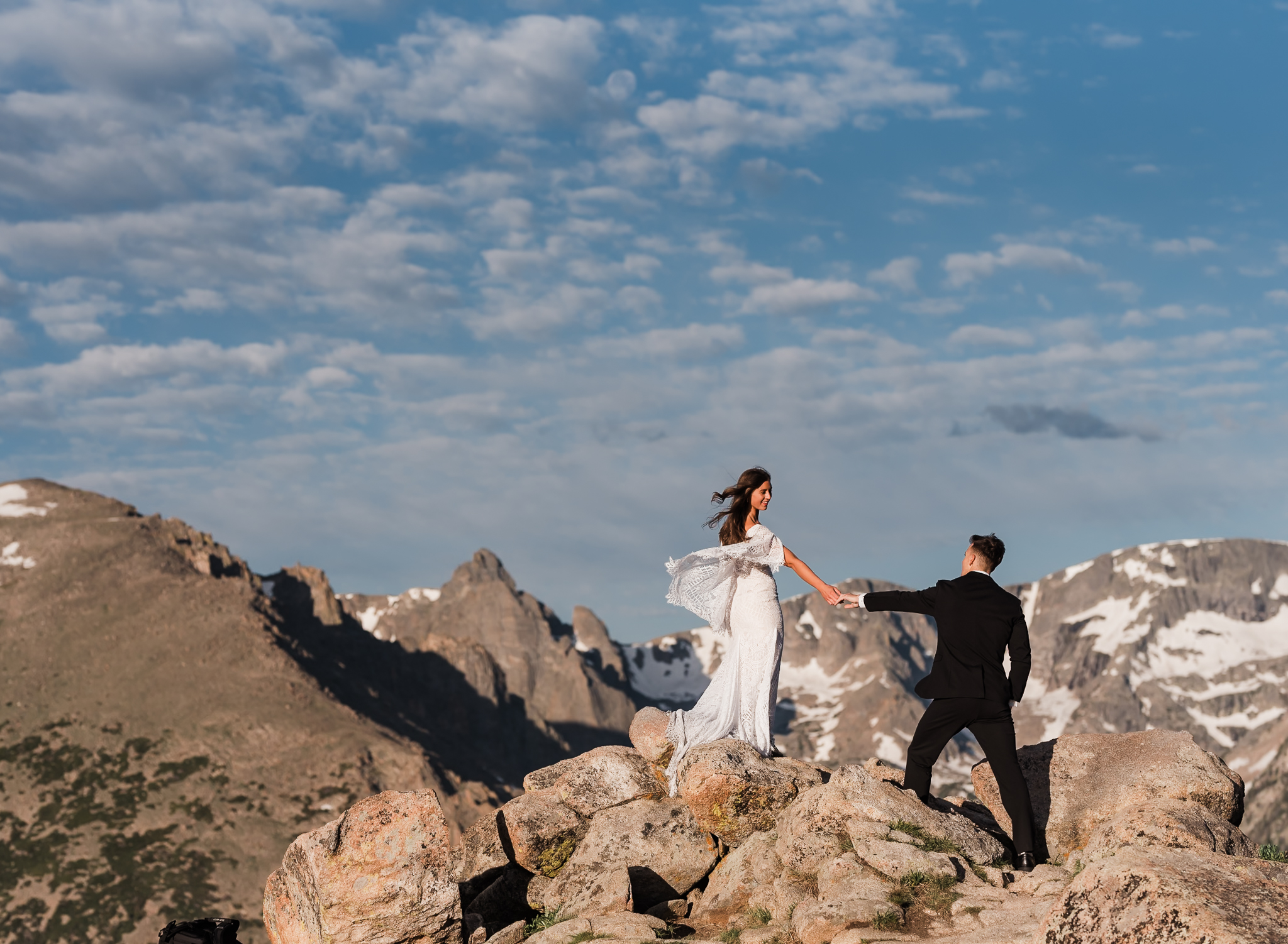 A bride stands on top of a rock holding her hand out to her groom with large mountains behind them at their Rocky Mountain National Park elopement