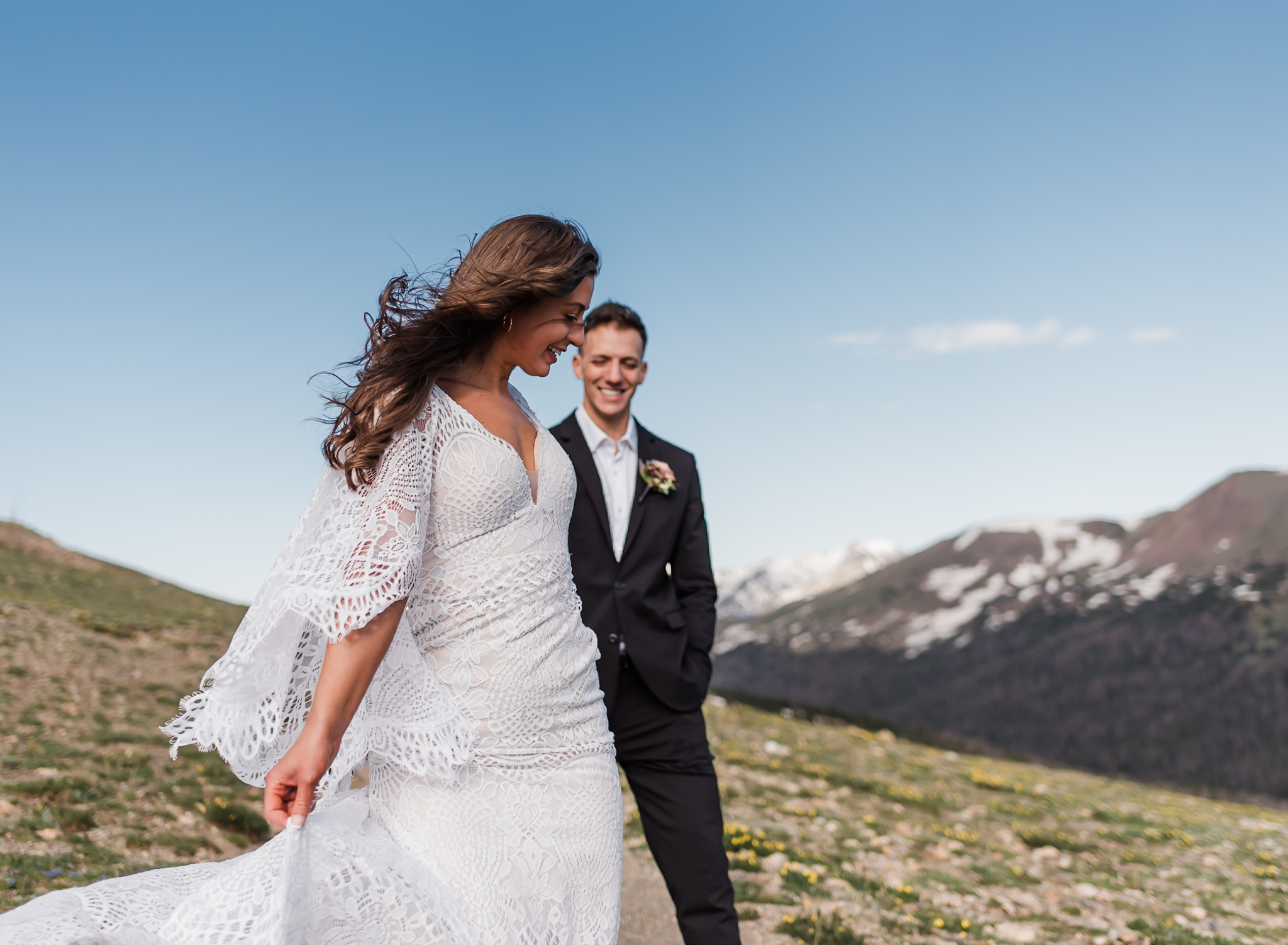 A bride and groom walk hand in hand with mountains behind them. The bride is leading the groom. 