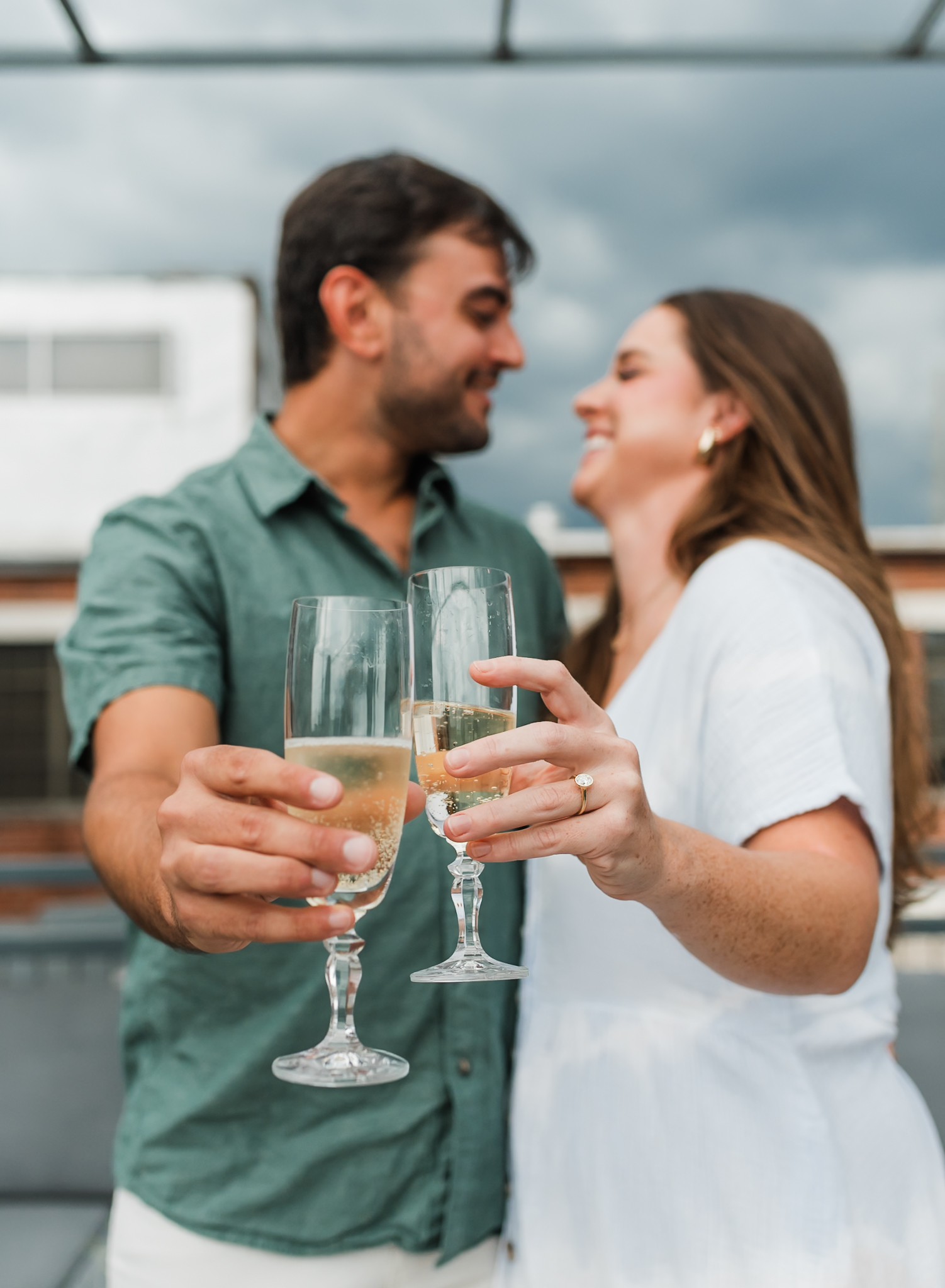 A man and woman toast with champagne glasses after getting engaged. The focus of the photo is on the woman's engagement ring and the champagne glasses.