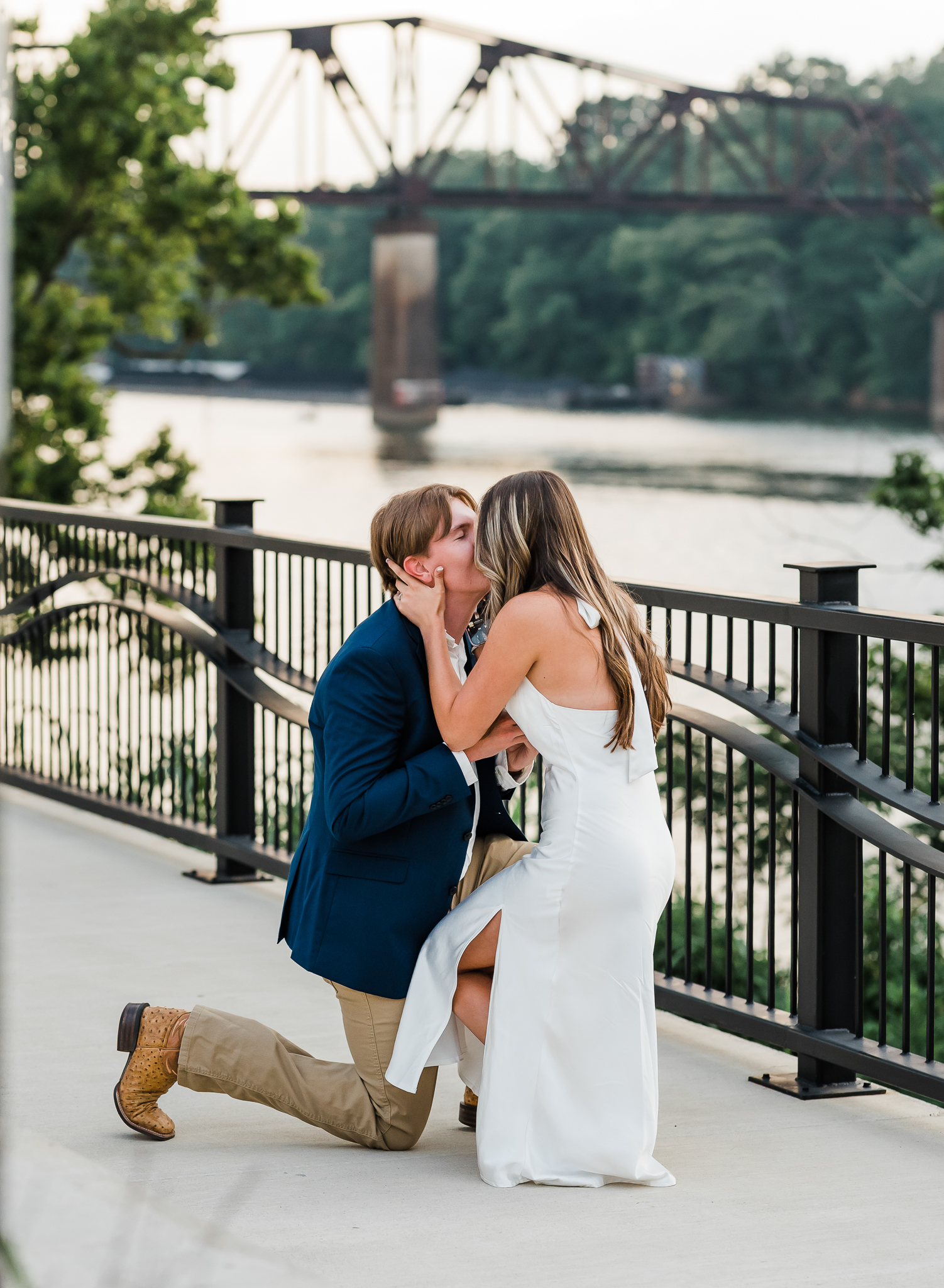 A man is down on one knee proposing to his girlfriend. The girl leans down on his level and kisses him.