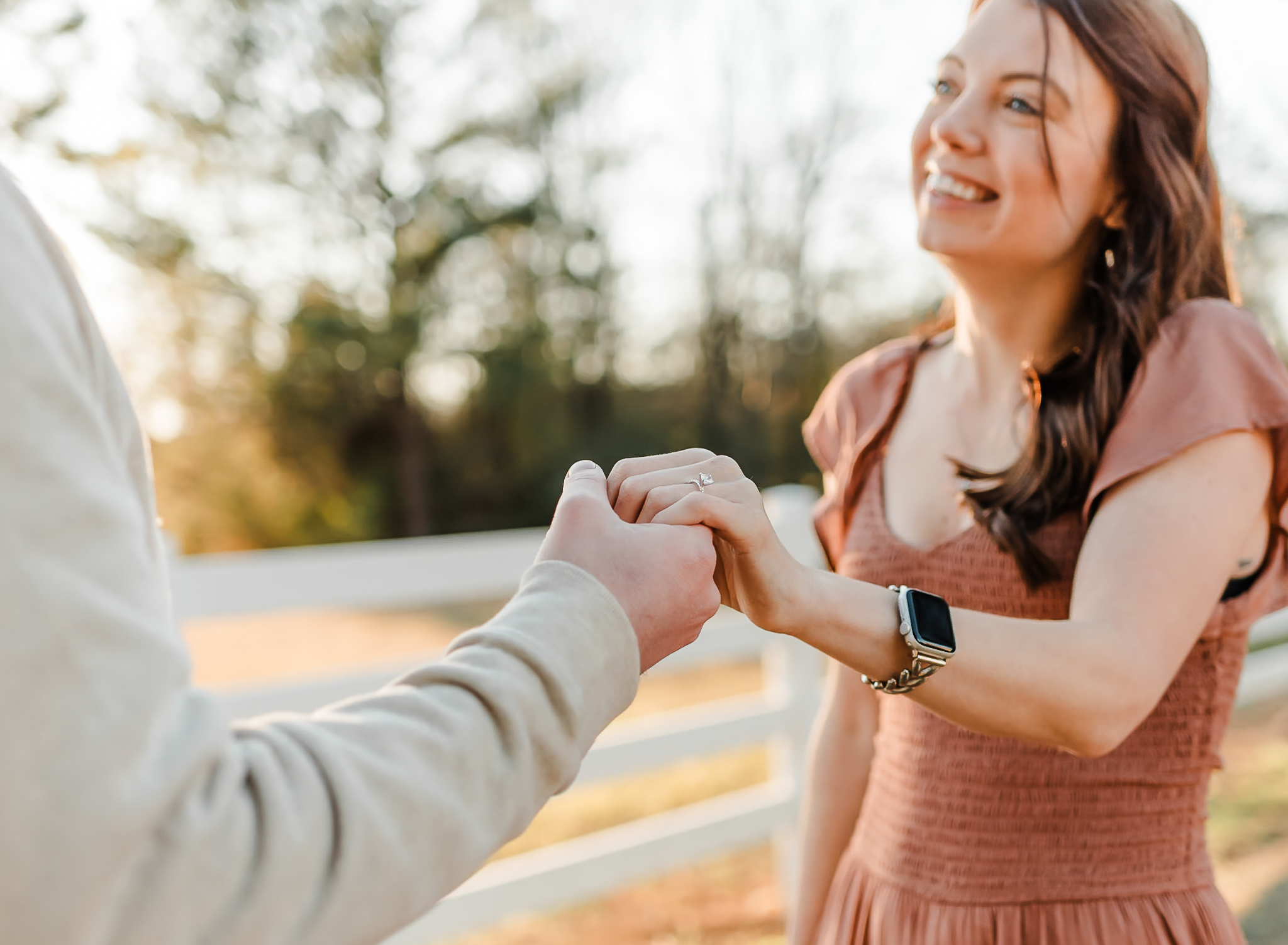 A man holding his fiances hand and smiling at her while she smiles back at him. The focus of the photo is on the engagement ring on the fiance's hand.