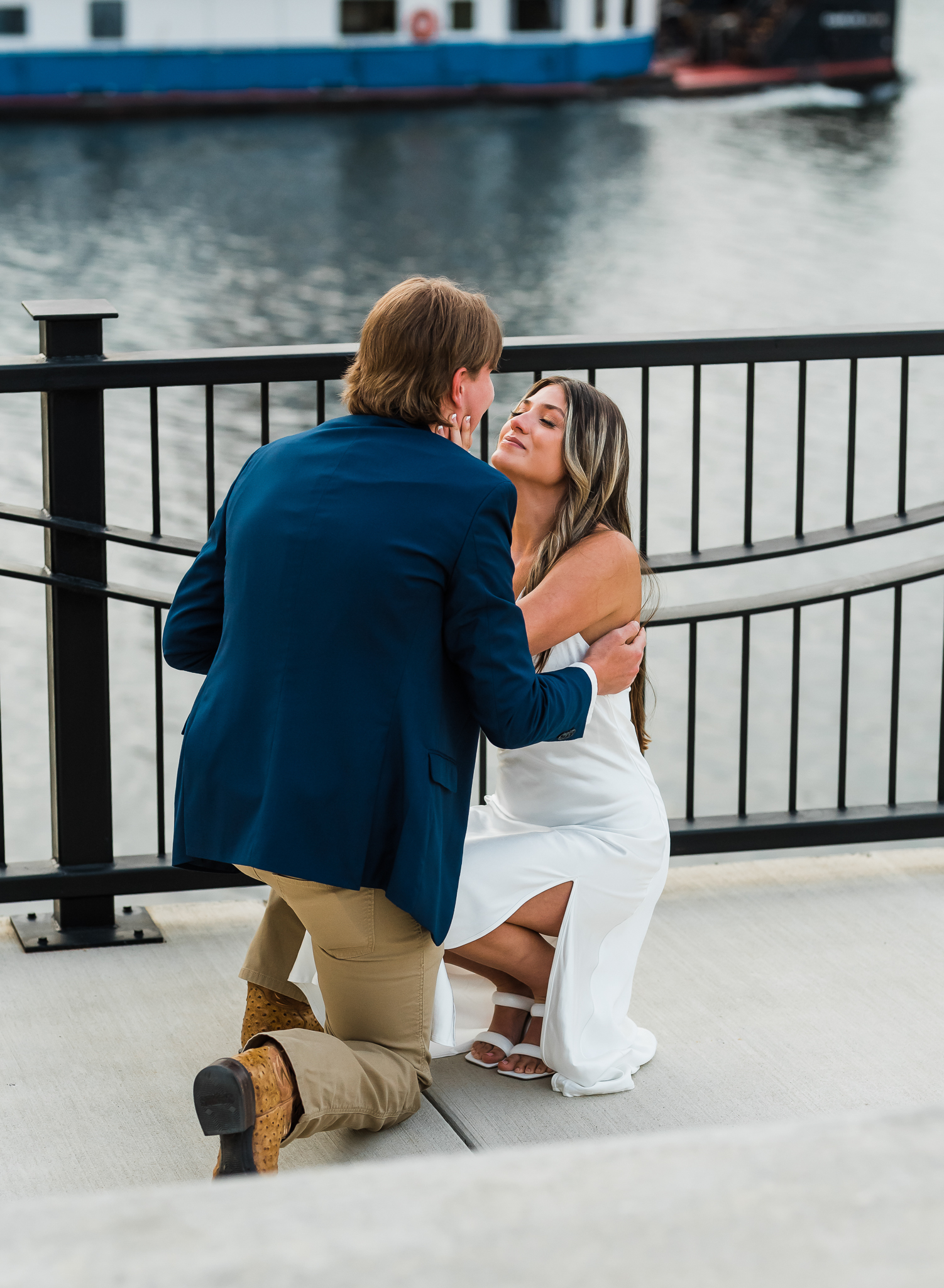 A man is down on one knee proposing to his girlfriend. The girl leans down on his level and holds his face while she is smilng and crying