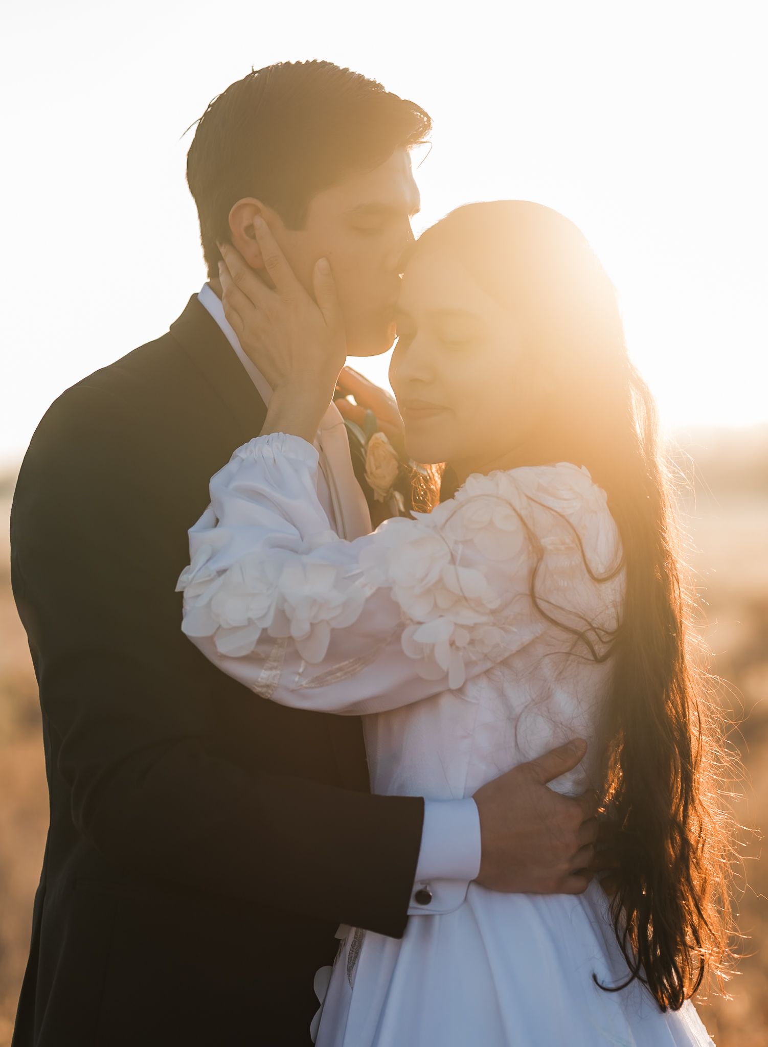 Sunrise elopement with a couple standing in golden hour light on a wide open plain. The groom is kissing the bride on the temple