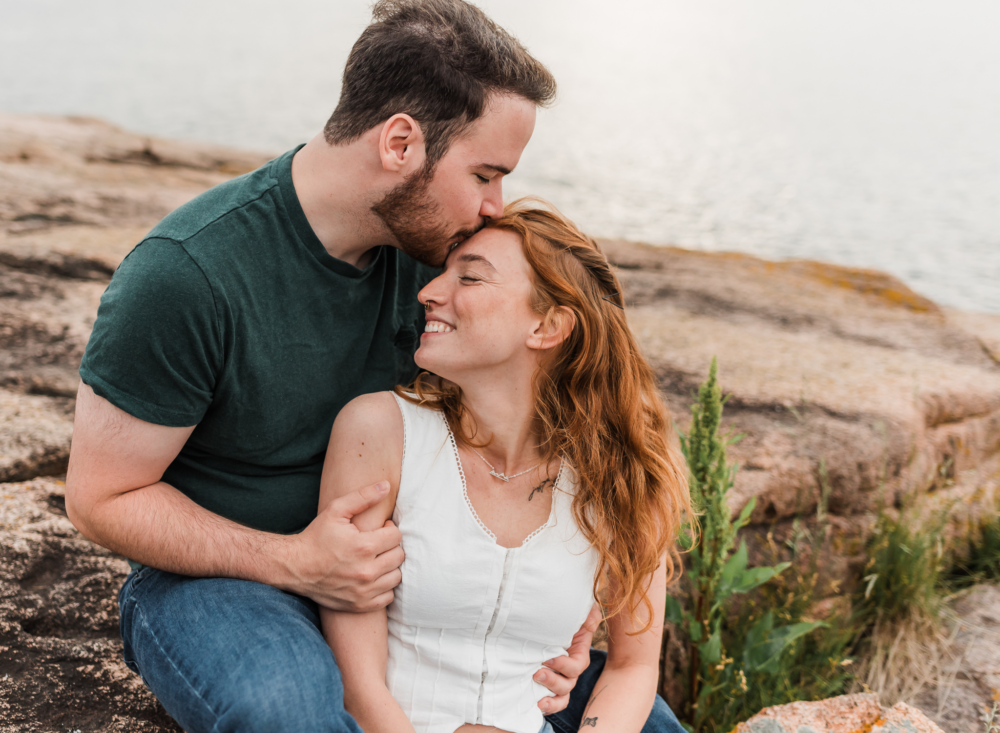A man and woman sit and snuggle up on the rocky coast of Acadia National Park in Maine