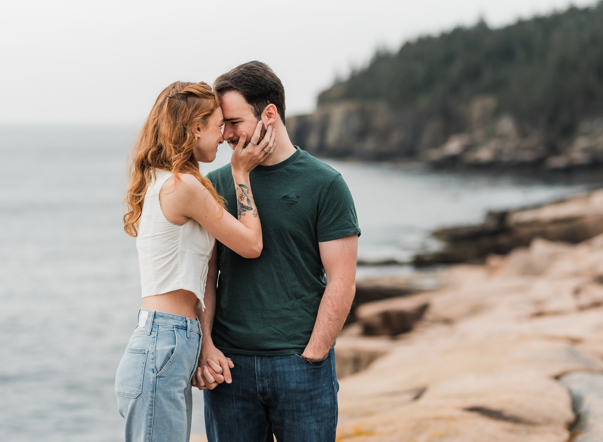 A woman and man stand forehead to forehead looking into each others eyes. The woman's hand is on the man's cheek. They are on the rocky coast of Acadia National Park in Maine.
