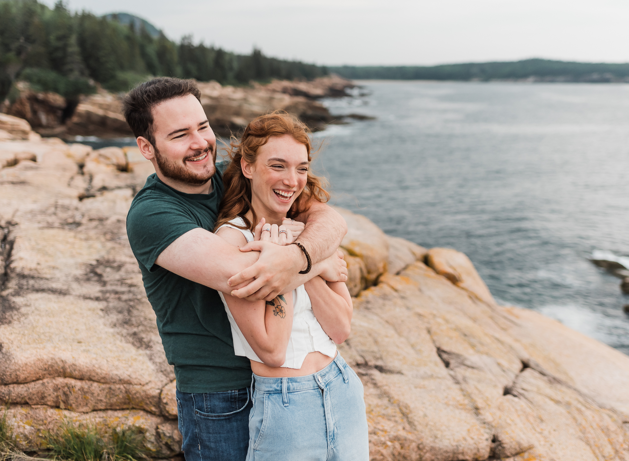 A man bear hugs a woman and they are laughing on the rocky coast of Acadia National Park in Maine