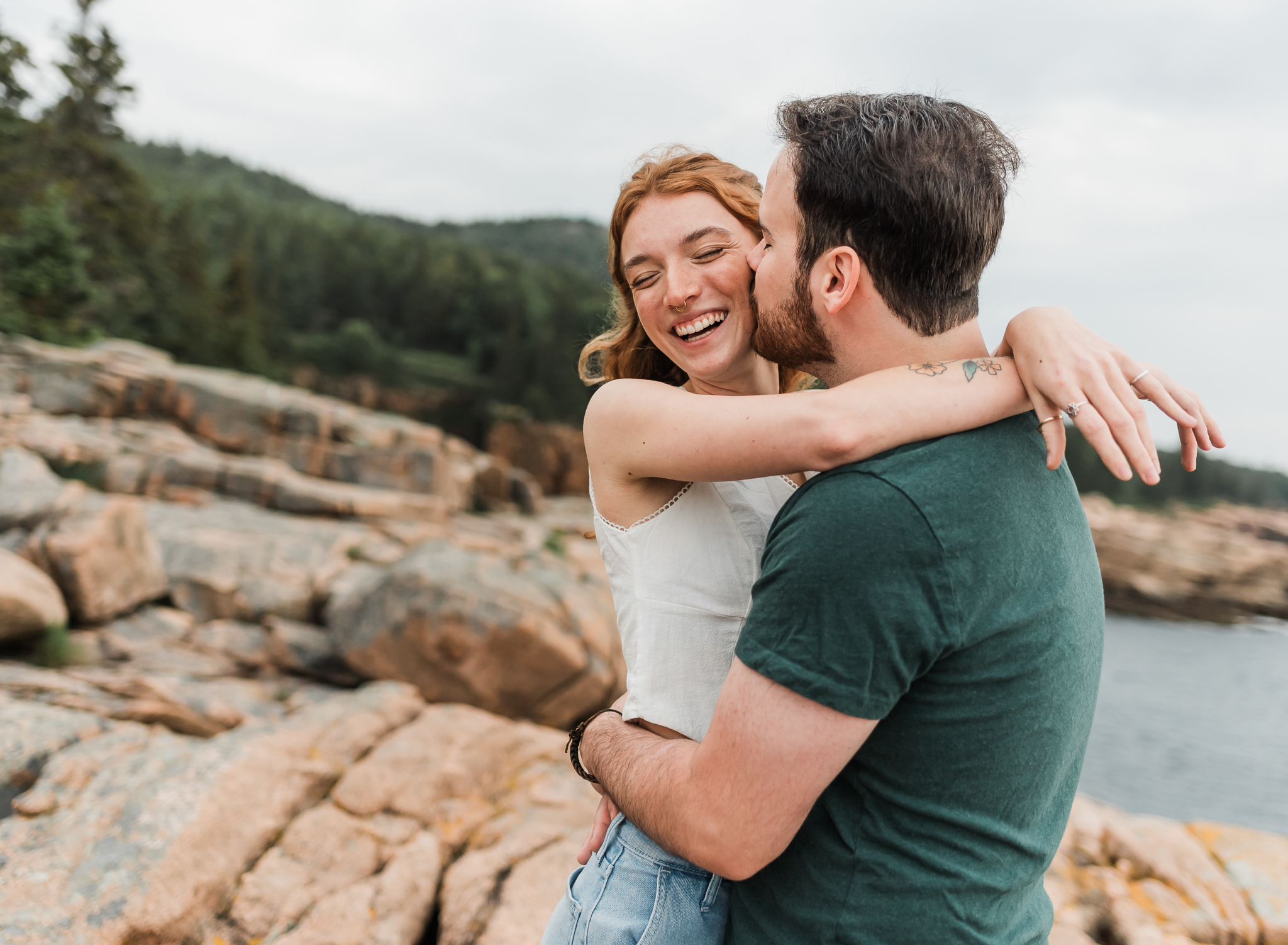 A man and woman hug and the man kisses the woman on the cheek on the rocky coast of Acadia National Park in Maine.