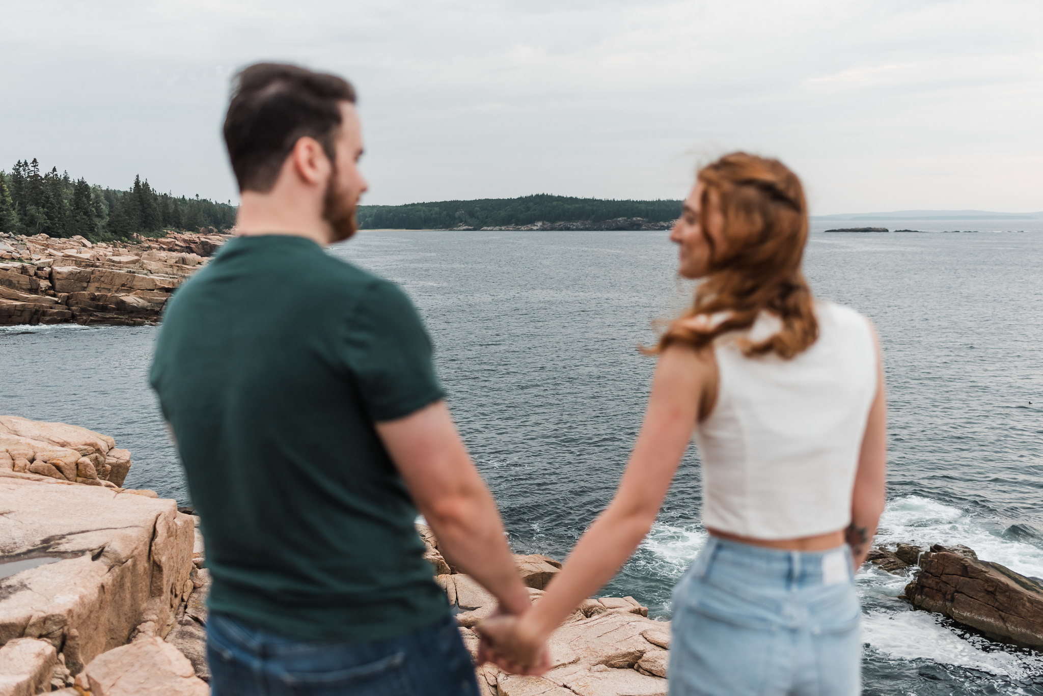 A man and woman hold hands and look at each other on the rocky coast of Acadia National Park in Maine. The image focus is on the landscape while the couple is out of focus.