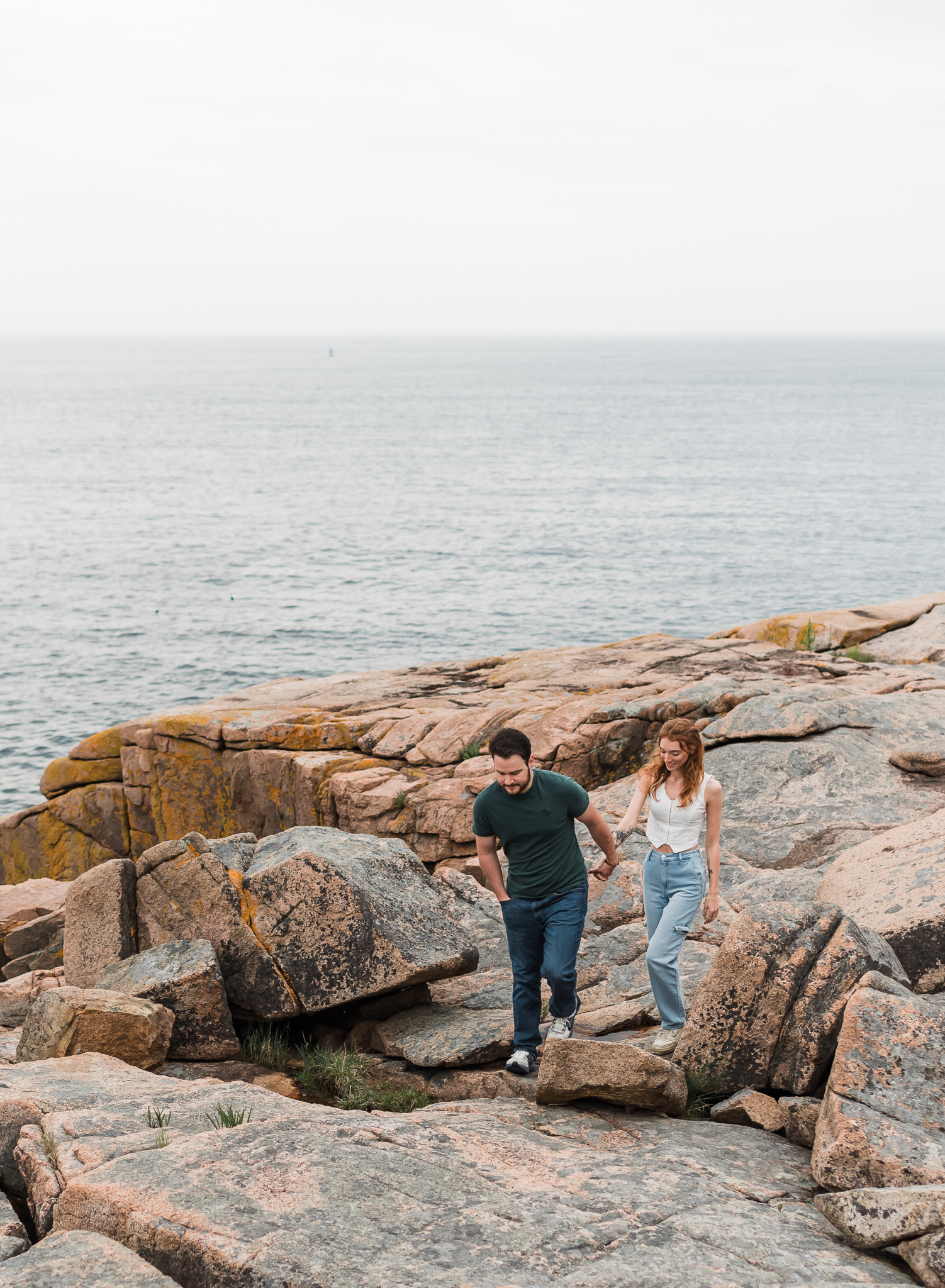 A man leads a woman walking hand in hand on the rocky coast of Acadia National Park in Maine