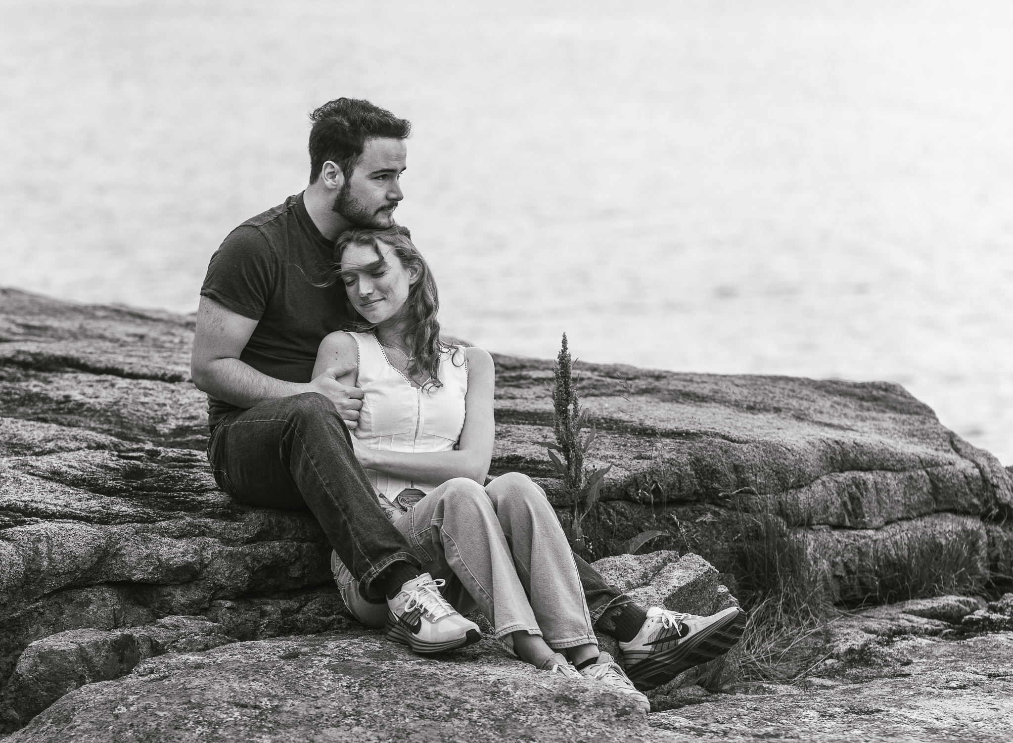 Man and woman sit and snuggle and look off into the distance on the rocky coast of Acadia National Park in Maine
