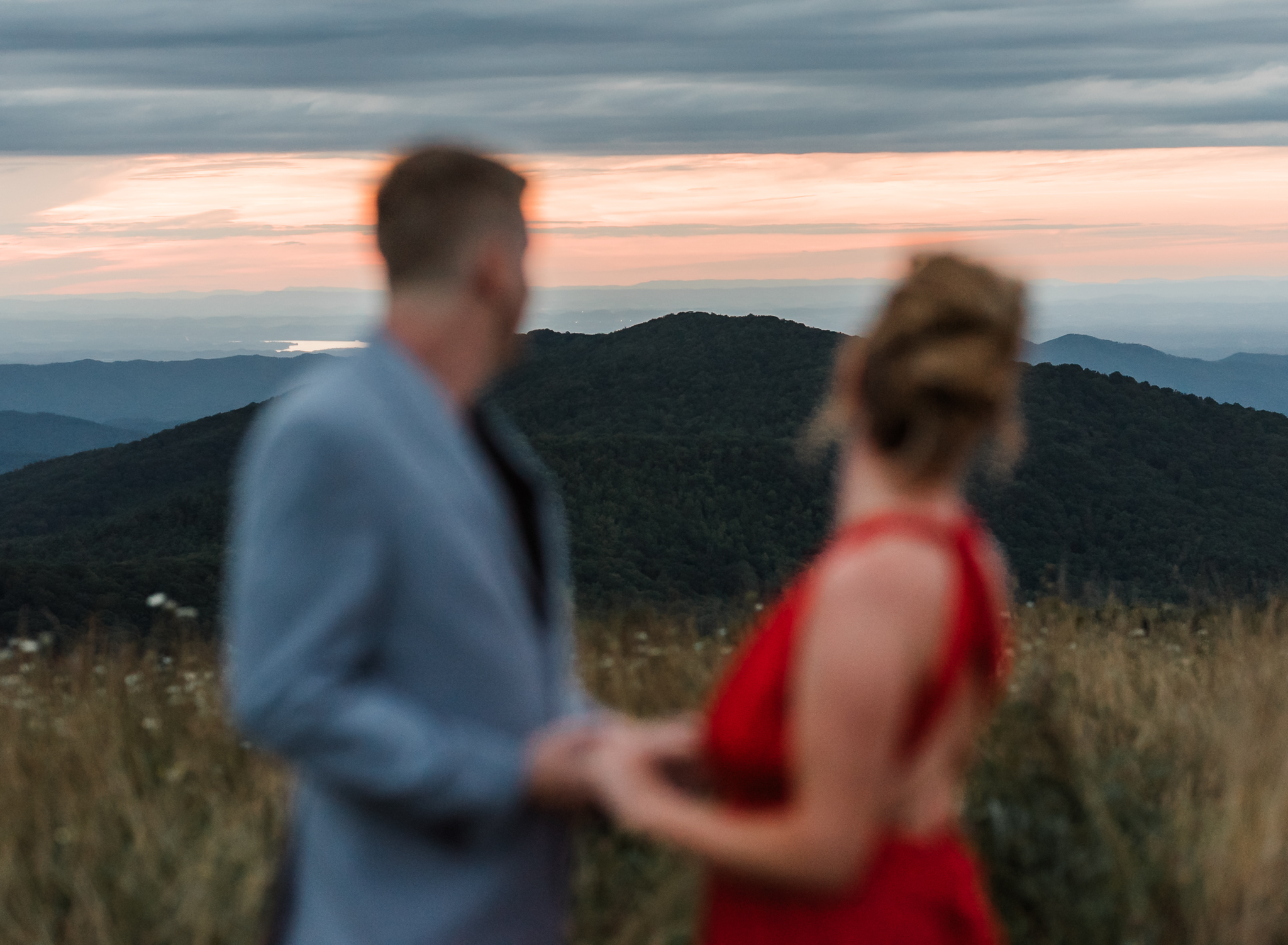 A man and woman look out at the colorful sky after the sun has set. The man and woman are intentionally blurry and the sky and mountains are in focus.