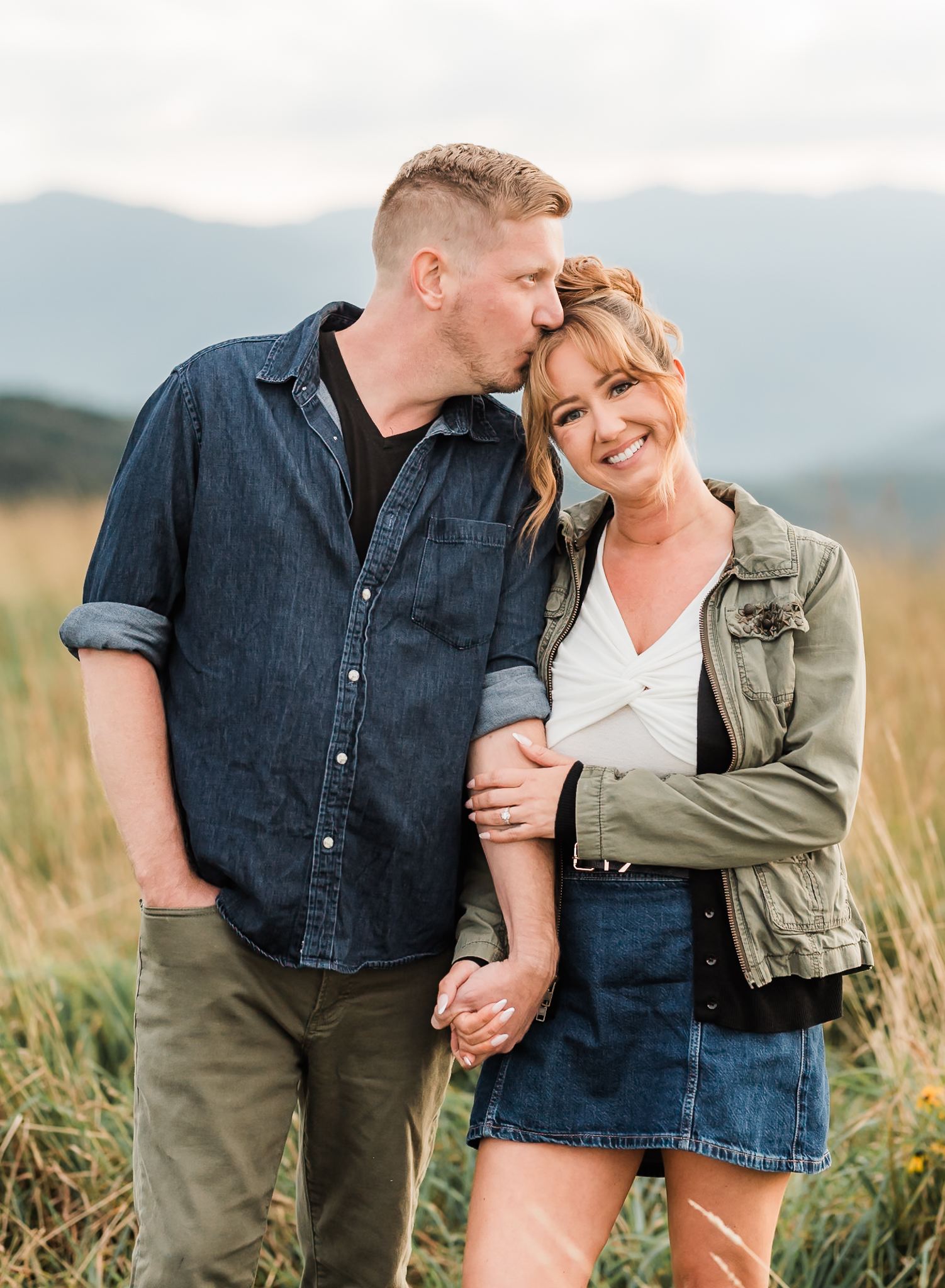 A man kisses his fiance on the temple while the woman smiles at the camera.